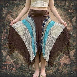 May include: A midi-length skirt featuring a wide brown waistband and a flared, handkerchief-style hem. The skirt is constructed from alternating vertical panels of solid brown fabric, teal patterned fabric, and white floral lace overlay.