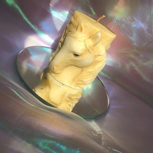 May include: A cream-colored horse-shaped candle with a detailed mane and face, resting on a heart-shaped mirror. The candle has a wick and sits against a shimmering, iridescent background with purple and green hues.