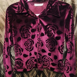 May include: A long-sleeved, button-down shirt in a deep magenta hue, adorned with a pattern of black roses. The shirt features a collared neckline and is made of a soft, velvety material. The garment is displayed on a white hanger.