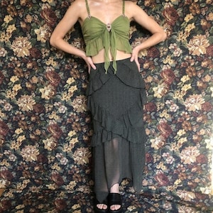 Op de afbeelding: Een groene crop top met juwelen bandjes en een strik detail, gecombineerd met een zwarte polkadot rok met gelaagde ruches. De outfit wordt gecompleteerd met zwarte plateausandalen, tegen een bloemenachtergrond.