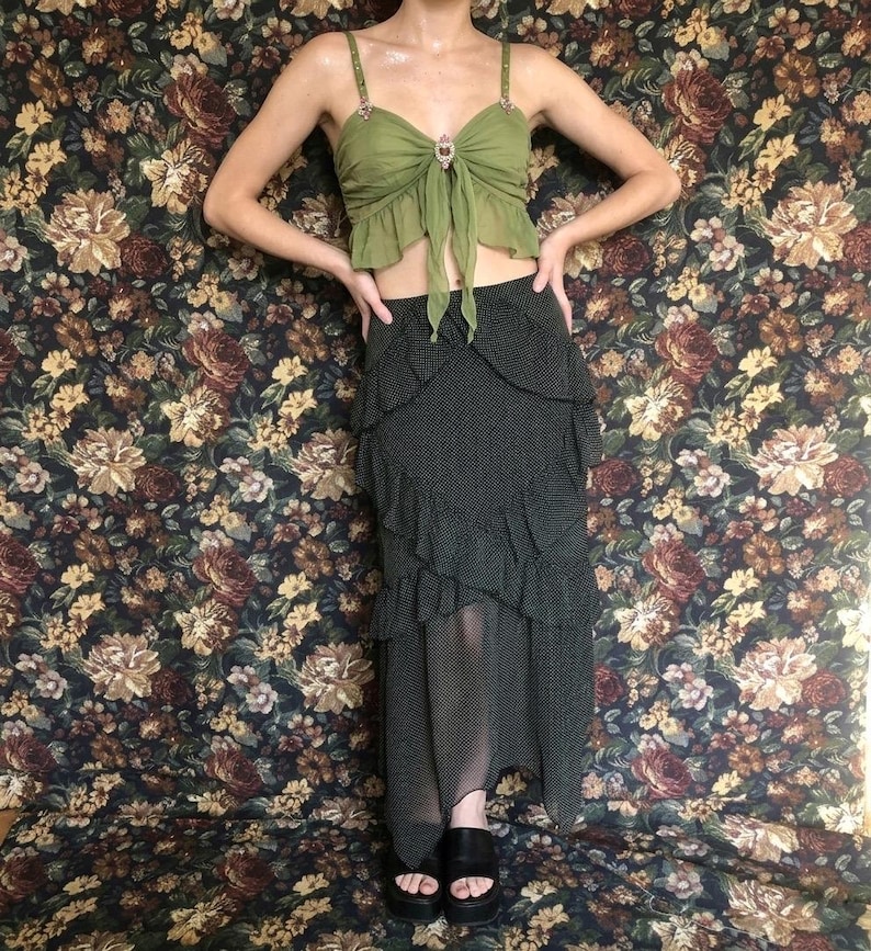 Op de afbeelding: Een groene crop top met juwelen bandjes en een strik detail, gecombineerd met een zwarte polkadot rok met gelaagde ruches. De outfit wordt gecompleteerd met zwarte plateausandalen, tegen een bloemenachtergrond.
