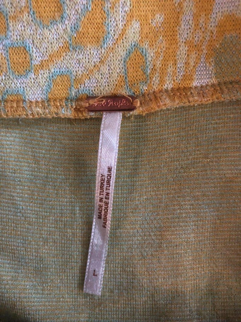 Pode incluir: Uma etiqueta branca com texto preto que diz "Made in Turkey Fabrique en Turquie" anexada a um tecido estampado amarelo e verde. A etiqueta tem um tamanho "L" impresso.