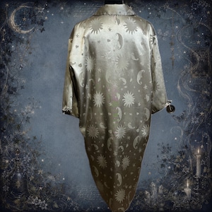 May include: A long-sleeved, button-down shirt in a muted gold tone featuring a repeating jacquard pattern of suns, crescent moons, and stars. The garment has a classic collar, a loose fit, and a smooth, slightly reflective fabric finish.