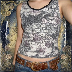 May include: A sleeveless tank top featuring a dark gray and white floral pattern with a black trim neckline, paired with blue denim jeans and a wide brown braided leather belt with a large ornate metal buckle and metallic stud accents.