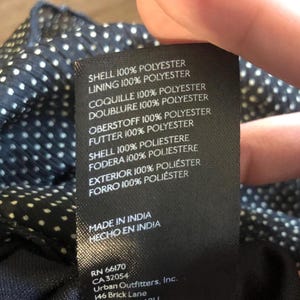 Op de afbeelding: Een zwart label met witte tekst, met details over de stof samenstelling van een kledingstuk. Het label vermeldt "SHELL 100% POLYESTER" en "LINING 100% POLYESTER". Het bevat ook "MADE IN INDIA" en het adres van Urban Outfitters, Inc.