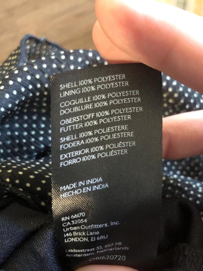 Op de afbeelding: Een zwart label met witte tekst, met details over de stof samenstelling van een kledingstuk. Het label vermeldt "SHELL 100% POLYESTER" en "LINING 100% POLYESTER". Het bevat ook "MADE IN INDIA" en het adres van Urban Outfitters, Inc.