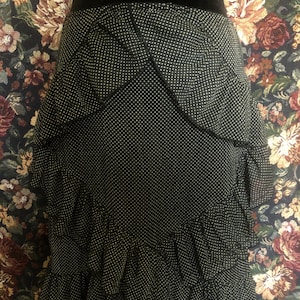 Op de afbeelding: Zwarte rok met witte stippen en gelaagde ruches. De rok heeft een hoge taille en een getailleerd silhouet. De stof lijkt een lichtgewicht materiaal te zijn, geschikt voor warmer weer. De rok is een klassiek en veelzijdig kledingstuk.