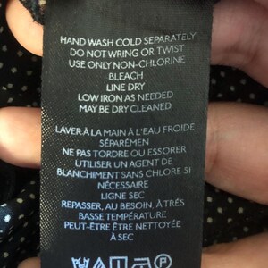 Op de afbeelding: Een zwart stoffen label met witte tekst met wasvoorschriften. Het label bevat zinnen als "Handwas koud apart," "Lijn drogen," en "Kan chemisch gereinigd worden." Er zijn ook verzorgingssymbolen aanwezig.