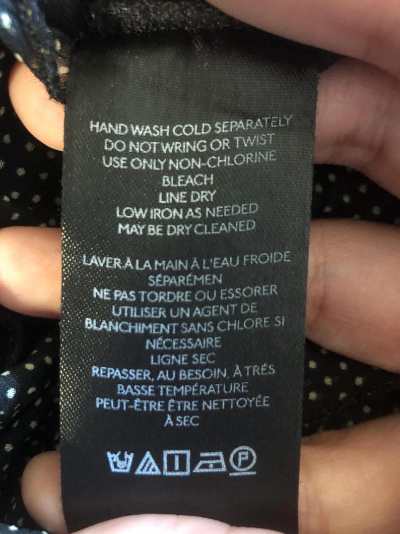 Op de afbeelding: Een zwart stoffen label met witte tekst met wasvoorschriften. Het label bevat zinnen als "Handwas koud apart," "Lijn drogen," en "Kan chemisch gereinigd worden." Er zijn ook verzorgingssymbolen aanwezig.