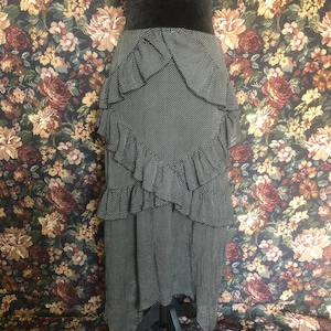 Op de afbeelding: Een zwart-witte polkadot rok met gelaagde ruches. De rok wordt getoond op een zwarte mannequin. De achtergrond heeft een bloemenpatroon met rozen en andere bloemen in tinten rood, bruin en geel.
