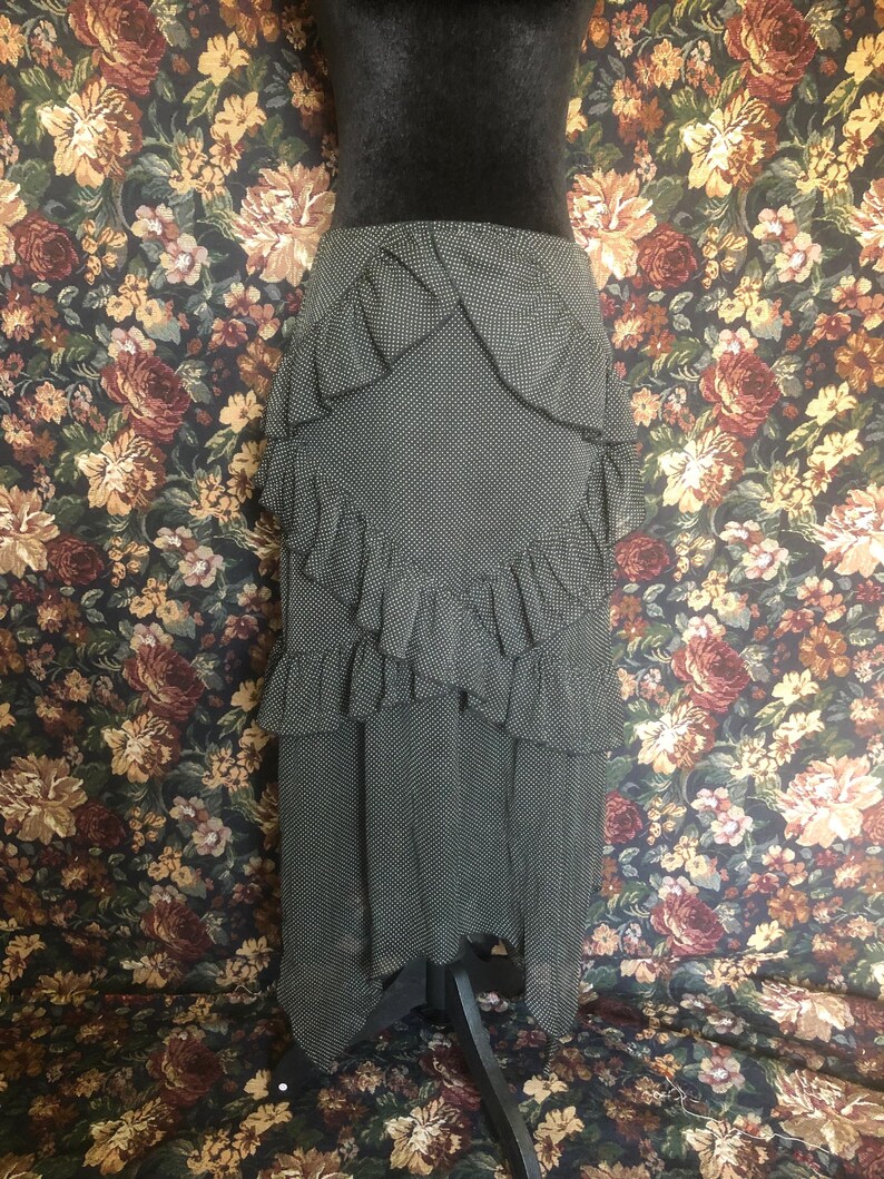 Op de afbeelding: Een zwart-witte polkadot rok met gelaagde ruches. De rok wordt getoond op een zwarte mannequin. De achtergrond heeft een bloemenpatroon met rozen en andere bloemen in tinten rood, bruin en geel.