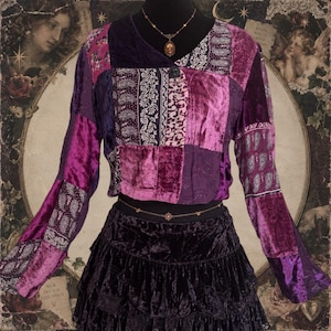 May include: A long-sleeve patchwork top featuring various shades of purple and magenta velvet and patterned fabric panels, paired with a black tiered velvet skirt. The top has a single button closure at the neckline and a V-shaped collar design.
