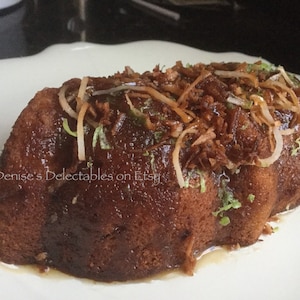 May include: A brown bundt cake topped with shredded coconut and a glaze. The cake is on a white plate. Denise's Delectables on Etsy is written on the cake.