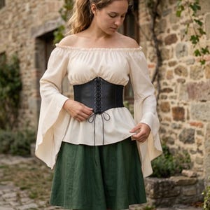 Könnte beinhalten: Eine cremefarbene Off-Shoulder-Bluse mit langen, ausgestellten Ärmeln. Ein schwarzes Korsett mit Schnürung vorne schnürt die Taille ein. Das Outfit wird mit einem grünen Rock kombiniert und kreiert einen mittelalterlichen oder Renaissance-inspirierten Look.