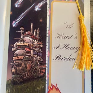 May include: Two decorative bookmarks. One shows a moving castle illustration with a night sky and shooting stars. The other has the text "A Heart's A Heavy Burden" and a cartoon fire character. Both have yellow tassels.