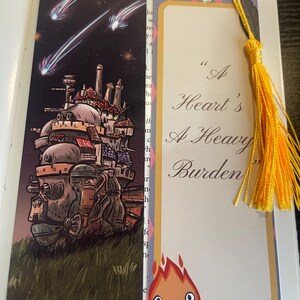 May include: Two decorative bookmarks with yellow tassels. One shows a moving castle under a starry sky. The other has the quote "A Heart's A Heavy Burden" and a cartoon flame character.