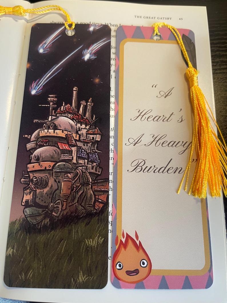 May include: Two decorative bookmarks with yellow tassels. One shows a moving castle under a starry sky. The other has the quote "A Heart's A Heavy Burden" and a cartoon flame character.