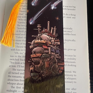 May include: Bookmark with a detailed illustration of a moving castle, set against a dark night sky with shooting stars. It has a yellow tassel attached to the top. The bookmark is made of card.
