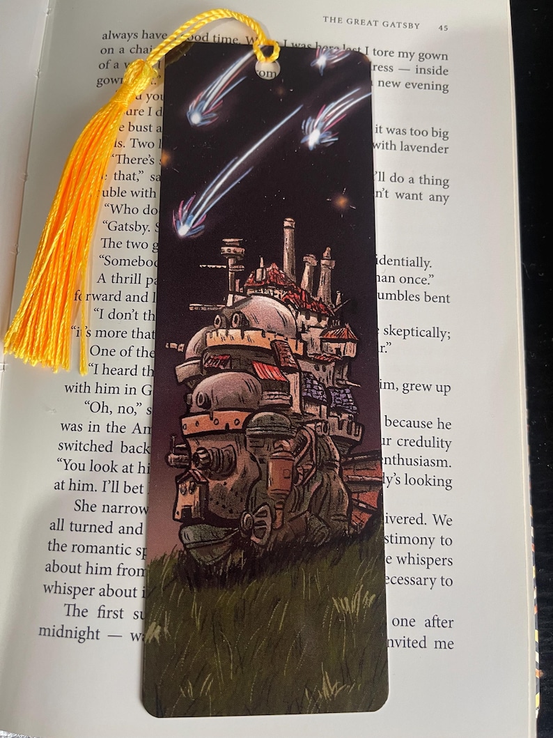 May include: Bookmark with a detailed illustration of a moving castle, set against a dark night sky with shooting stars. It has a yellow tassel attached to the top. The bookmark is made of card.
