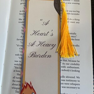 May include: A bookmark with the quote "A Heart's A Heavy Burden" in a decorative font. The bookmark has a cream background, a patterned border, and a cartoon flame character. A yellow tassel is attached at the top.