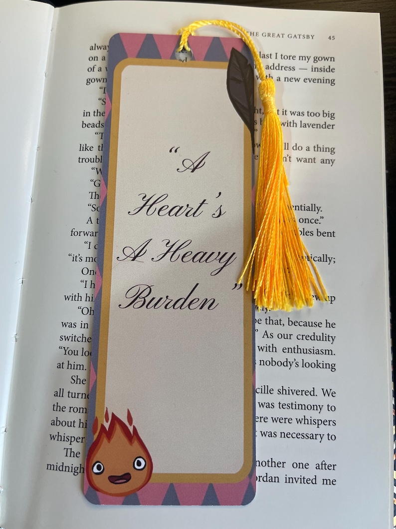 May include: A bookmark with the quote "A Heart's A Heavy Burden" in a decorative font. The bookmark has a cream background, a patterned border, and a cartoon flame character. A yellow tassel is attached at the top.