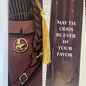 May include: Two bookmarks with a dark brown background. The bookmark on the left features a stylized illustration of a mockingjay pin with a gold finish. The bookmark on the right features a forest scene with the text "May the odds be ever in your favor".