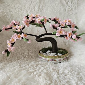 May include: A crocheted bonsai tree with pink flowers and green leaves in a white ceramic pot with a floral design.
