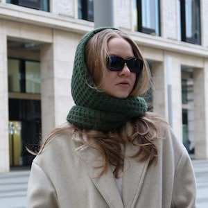 May include: A woman wearing a dark green knitted hood and a light beige coat. The hood is wrapped around the head and neck. The woman is also wearing black sunglasses. The background is a building with large windows.