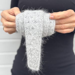 Könnte beinhalten: Ein hellgrauer Strickartikel mit strukturiertem Muster und einem flauschigen, gefransten Rand. Der Artikel wird von einer Person gehalten, die ein schwarzes Oberteil trägt. Der Strickartikel scheint ein Schal oder ein ähnliches Accessoire zu sein.