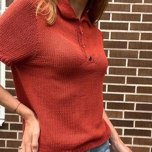 May include: A rust-colored knitted polo shirt with short sleeves and a collared neckline. The shirt has a buttoned placket and a textured knit pattern. The garment is made of a soft, breathable material, perfect for casual wear.