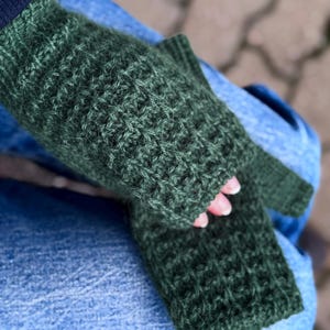 Könnte beinhalten: Dunkelgrüne Strick-Fingerhandschuhe mit strukturiertem Muster. Die Handschuhe sind aus einem weichen, warmen Material gefertigt. Sie werden auf einer Person gezeigt, die eine blaue Jeans trägt. Ideal für kalte Tage.