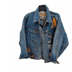 May include: A blue denim jacket featuring a large yellow letter A patch on the left chest, multiple smaller decorative patches, a button-up front, two chest pockets, and a tan leather-look brand label on the inside collar reading Gasoline.