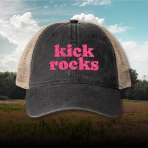 Könnte beinhalten: Schwarze Trucker-Cap mit dem pinkfarbenen Schriftzug "kick rocks". Die Kappe hat einen Mesh-Rücken und einen gebogenen Schirm. Der Hintergrund zeigt ein Feld und den Himmel.