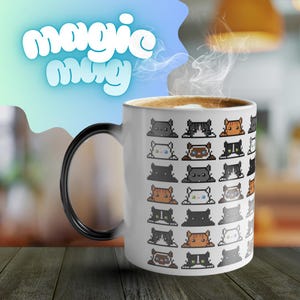 Puede incluir: Una taza de cerámica blanca con asa negra, llena de café y vapor. La taza está decorada con caras de gatos pixeladas en varios colores. Las palabras "magic mug" están escritas en una fuente estilizada.