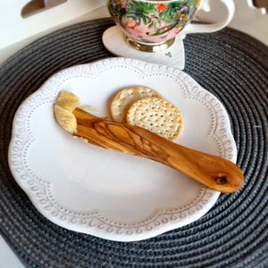 May include: A white ceramic plate with a scalloped edge and a decorative pattern. A wooden butter knife with a spread of butter on it is on the plate. Two round crackers are also on the plate.