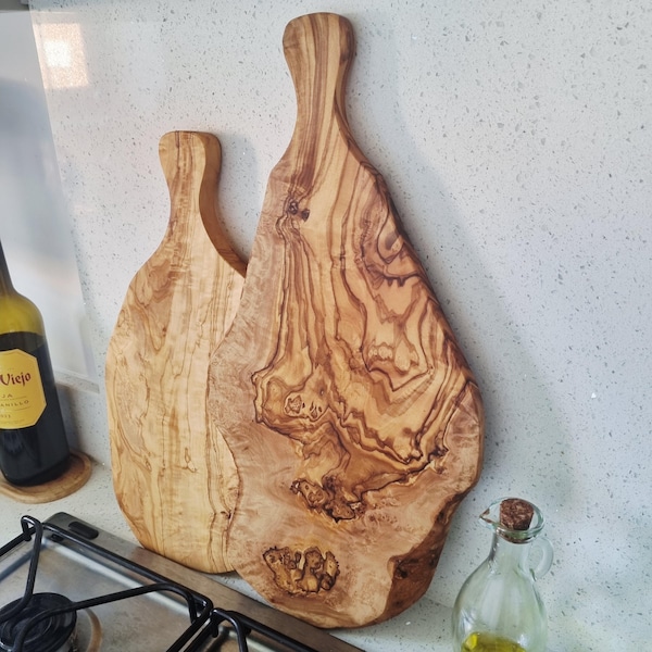 Op de afbeelding: Twee houten snijplanken met opvallende natuurlijke houtnerven en noesten, voorzien van langwerpige handvatten. De planken zijn gemaakt van lichtbruin hout met donkerdere, wervelende texturen en staan rechtop naast een wijnfles en een oliekaraf.