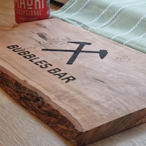 May include: A wooden serving board with the text "BUBBLES BAR" and a graphic of a crossed hammer and pickaxe. The board has a natural edge and a light brown color. A red can is visible in the background.