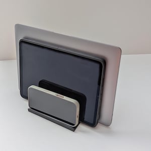 May include: A black metal stand holding a silver laptop, a black tablet, and a gray smartphone. The devices are stacked, with the laptop in the back and the phone in the front. The stand is on a white surface.