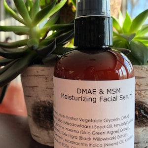 Puede incluir: Una botella de vidrio marr&oacute;n con una etiqueta blanca que dice "DMAE & MSM Suero Facial Hidratante". La botella tiene una tapa de bomba negra y contiene 118 ml de suero.
