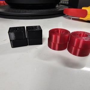May include: Two stacks of black, square-shaped objects and two rolls of red, ribbon-like material are arranged on a white surface. The black objects appear to be stacked blocks. The red rolls have a circular shape.