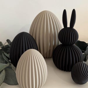 Könnte beinhalten: Eine Sammlung dekorativer Objekte zum Thema Ostern. Die Anordnung umfasst gerippte, eiförmige Skulpturen in Schwarz und Beige sowie eine schwarze Hasenfigur. Die Objekte sind auf einer weißen Oberfläche mit grünen Blättern angeordnet.