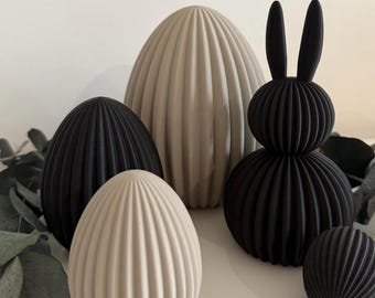 May include: A collection of decorative Easter-themed objects. The arrangement includes black and beige ribbed egg-shaped sculptures and a black bunny figure. The objects are arranged on a white surface with green foliage accents, creating a minimalist aesthetic.