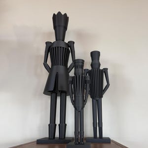 Könnte beinhalten: Drei schwarze dekorative Skulpturen von stilisierten Figuren. Die größte Figur hat ein kronenartiges Kopfstück und einen Rock. Die anderen beiden Figuren sind kleiner und haben zylindrische Körper mit vertikalen Linien. Alle Figuren stehen auf rechteckigen Sockeln.
