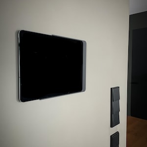 Op de afbeelding: Een zwart tablet met zilveren frame, gemonteerd op een lichtbeige muur. Het scherm van de tablet is zwart. Zwarte lichtschakelaars zijn rechts van de tablet te zien. De muur heeft een gladde afwerking, wat een moderne, minimalistische uitstraling creëert.