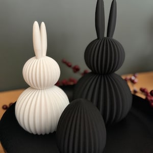 Könnte beinhalten: Zwei dekorative Hasenfiguren, eine weiße und eine schwarze, mit einem passenden schwarzen Ei. Die Hasen haben einen gerippten, kugelförmigen Körper und lange, schlanke Ohren. Die Objekte werden auf einer schwarzen Oberfläche präsentiert.