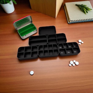 May include: An assortment of black pill organizers and a small metal container with the word "MOEIA" printed on the lid. The organizers are rectangular with multiple compartments, some labeled with days of the week. Several white pills are scattered on the wooden surface.