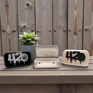May include: An assortment of small, rectangular trays with lids. Two trays feature the number "420" with a dripping effect and a cannabis leaf. One tray is black, the other is off-white. A silver tray has the word "VINOIDS" on the lid.
