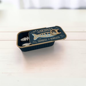 May include: A dark blue rectangular container with a lid featuring a tan pull tab and the text "not SARDINES" and "SKINLESS + BONELESS" above a light blue fish skeleton graphic. The container holds a white fish skeleton-shaped object inside.