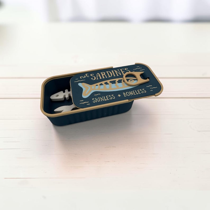 May include: A dark blue rectangular container with a lid featuring a tan pull tab and the text "not SARDINES" and "SKINLESS + BONELESS" above a light blue fish skeleton graphic. The container holds a white fish skeleton-shaped object inside.