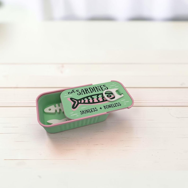 May include: A rectangular mint green tin container with a lid featuring a pink and black fish graphic, a white pull tab, and the text "not SARDINES" and "SKINLESS + BONELESS". Inside the container, two white fish-shaped objects are visible.